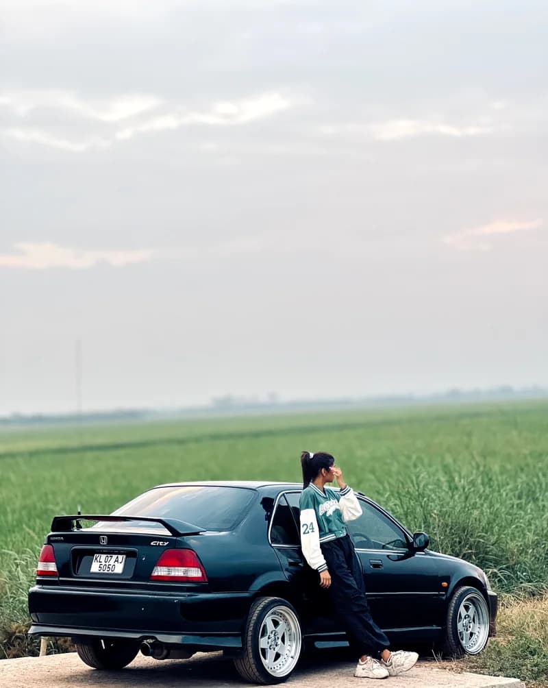 Closer source-photo crop of a dark Honda City and owner beside a green field.